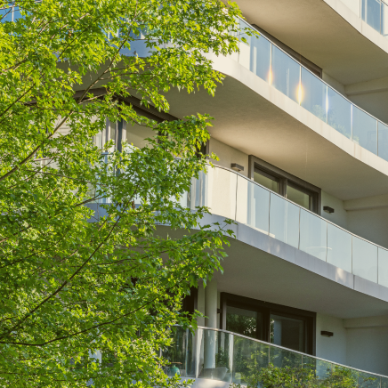 Modern residential building with curved glass balconies partially framed by green trees.
