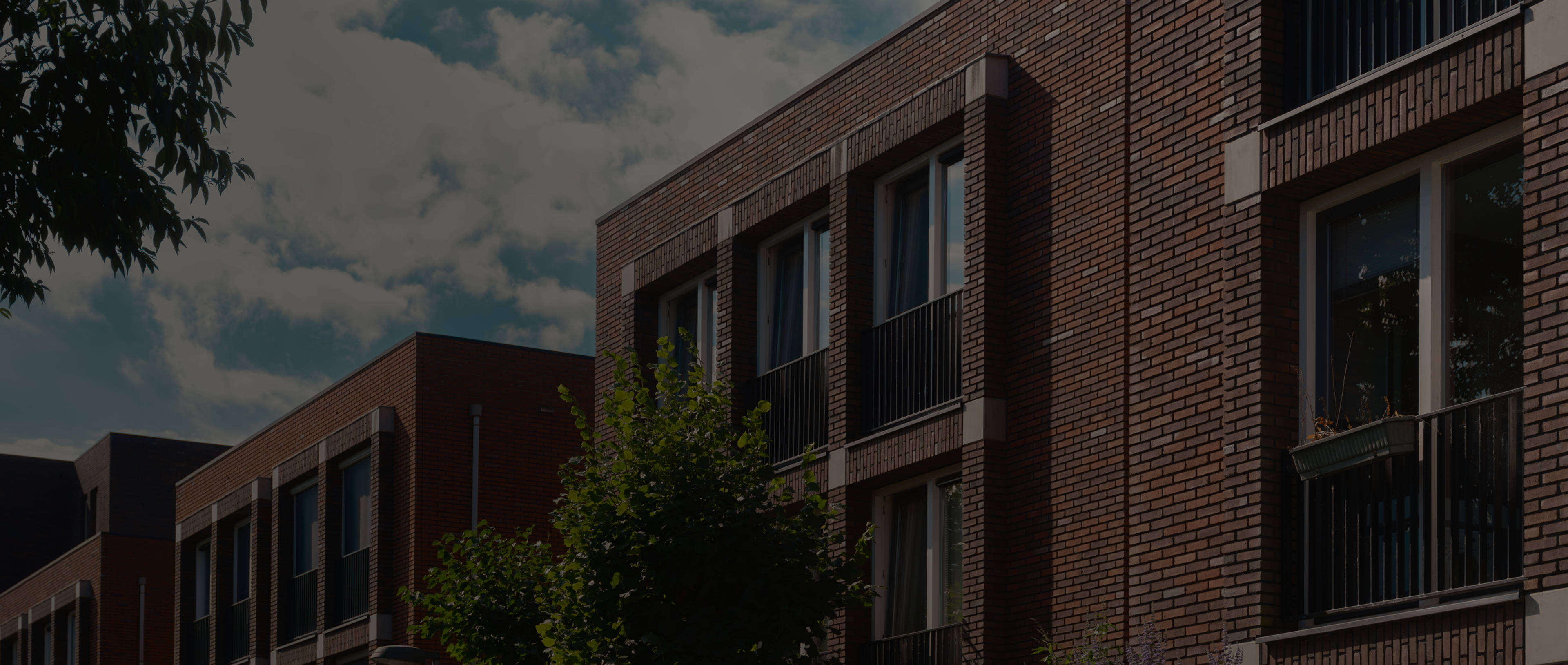 Modern brick residential buildings with balconies and trees beneath a partly cloudy sky.