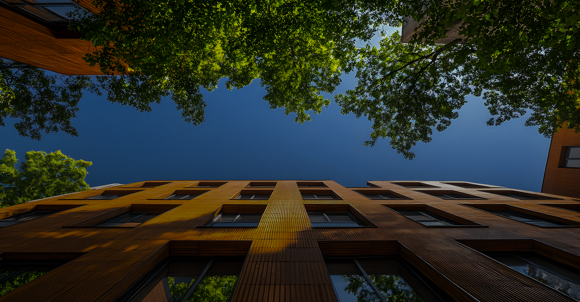 Low-angle view of modern residential buildings framed by leafy green trees and a deep blue sky.